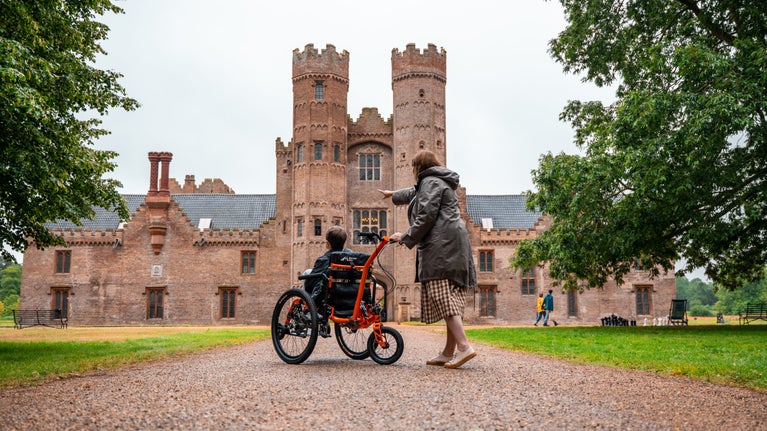 A woman points at Oxburgh Gatehouse and a young visitor sits in all terrain wheelchair looking